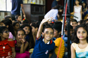 oval image. Young Indian boy in a navy blue, short-sleeved polo shirt has his hand up. Seated in a group of children, all wearing brightly coloured clothes.